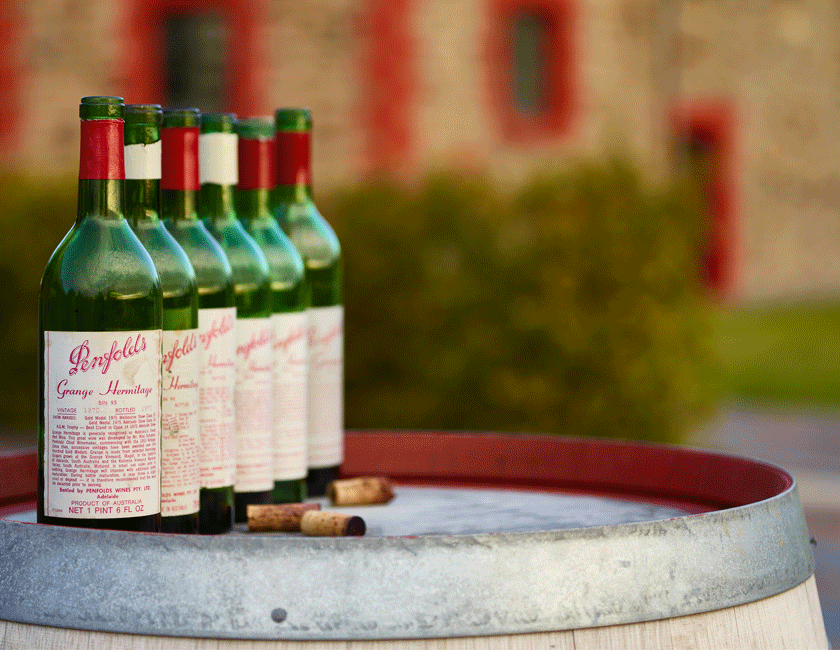 Six heritage Grange wine bottles from the 1970s sit outside on a barrel.  The cork sits beside and a heritage stone building is visible behind