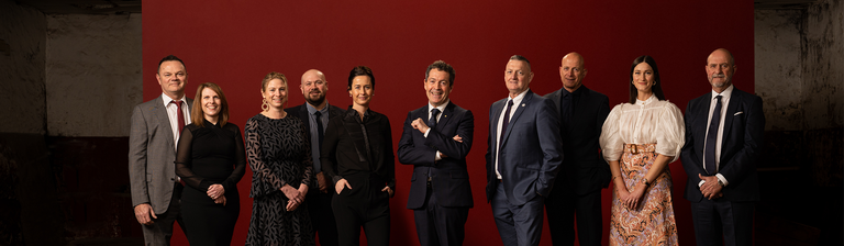 The Penfolds Winemaking Team.  10 people stand with red background behind. 