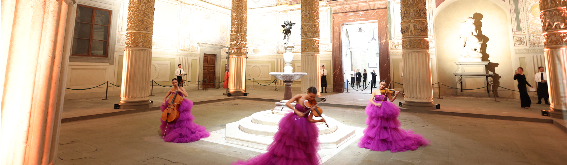 People in pink ball gowns play string instruments in a grand foyer