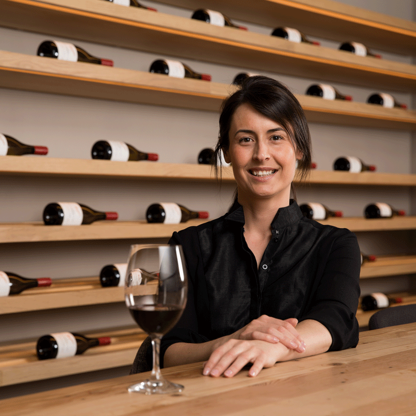 Steph Dutton, Penfolds Senior Winemaker, sits at a light wood table with rows of wine bottles behind her. 