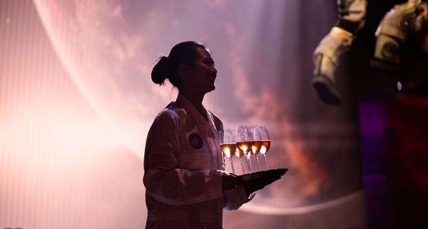 Waitress in space boiler suit greens guests with tray of champagne glasses