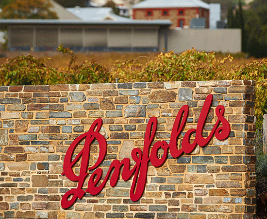 Signage. Red Penfolds logo against light yellow bricks with vines behind