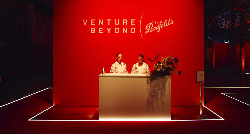 Two women greet guests at an entrance desk with red board behind. Text on board: Venture Beyond by Penfolds