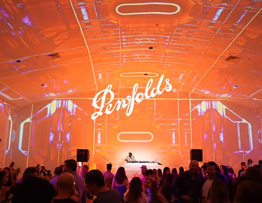 Photo across a busy event room  DJ stands on stage with orange projections covering the walls and ceiling above.  White Penfolds logo centred above the DJ booth.