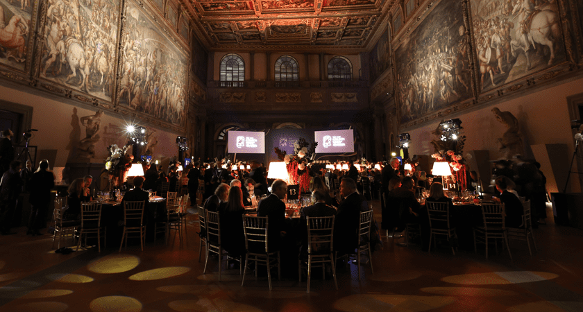 Black tie gala dinner scene.  Multiple round dining tables set against dramatic high ceilings with large historic paintings