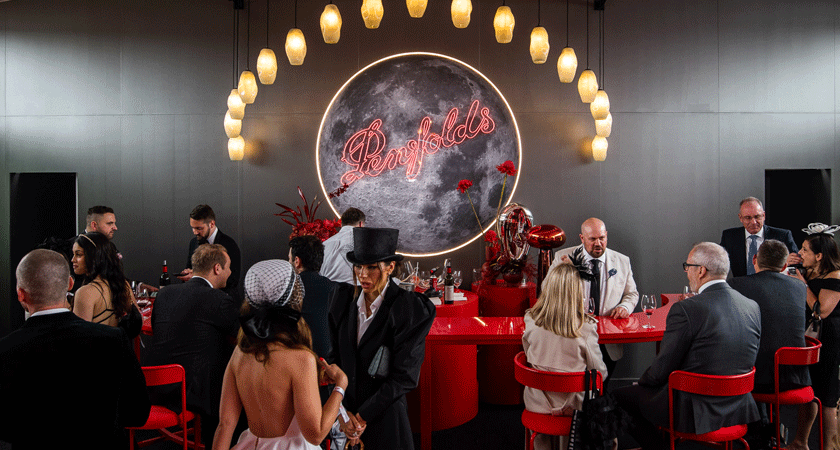 A black bar in the marquee with a neon Penfolds logo sign.  People mingle in front