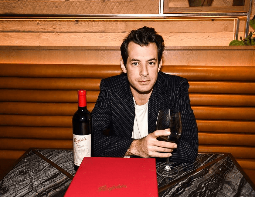 Mark Ronson sits at a table with a glass of red wine in hand.  Grange bottle sits on table