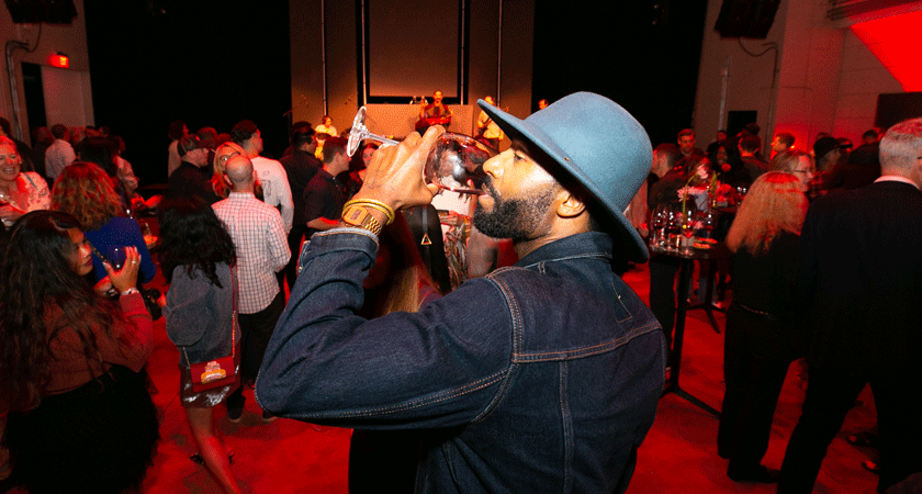 Guest poses at Venture Beyond event in LA.  He wears a blue felt hat and a denim jacket.  it's a profile view and he is sipping red wine
