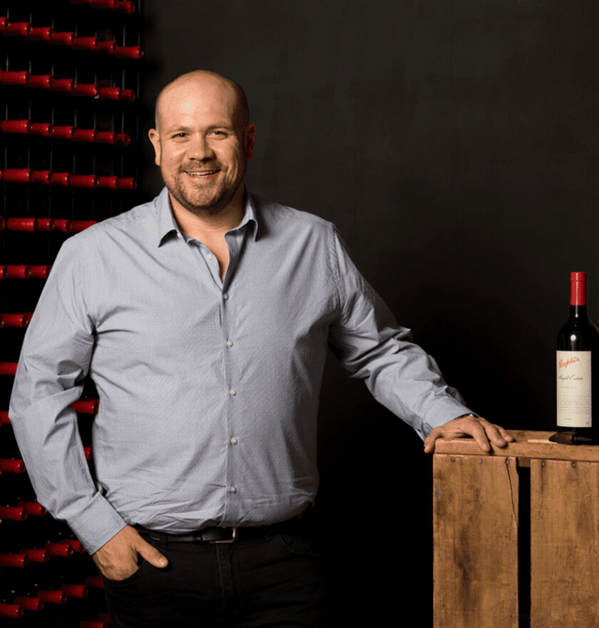 Matt Woo, Penfolds Winemaker, stands with a bottle of wine beside him and rows of red Penfolds bottles behind