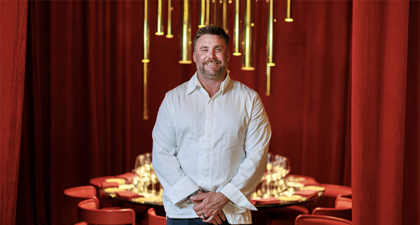 Chef, Scott Huggins, stands in AO restaurant with red curtains and a round red dining table behind